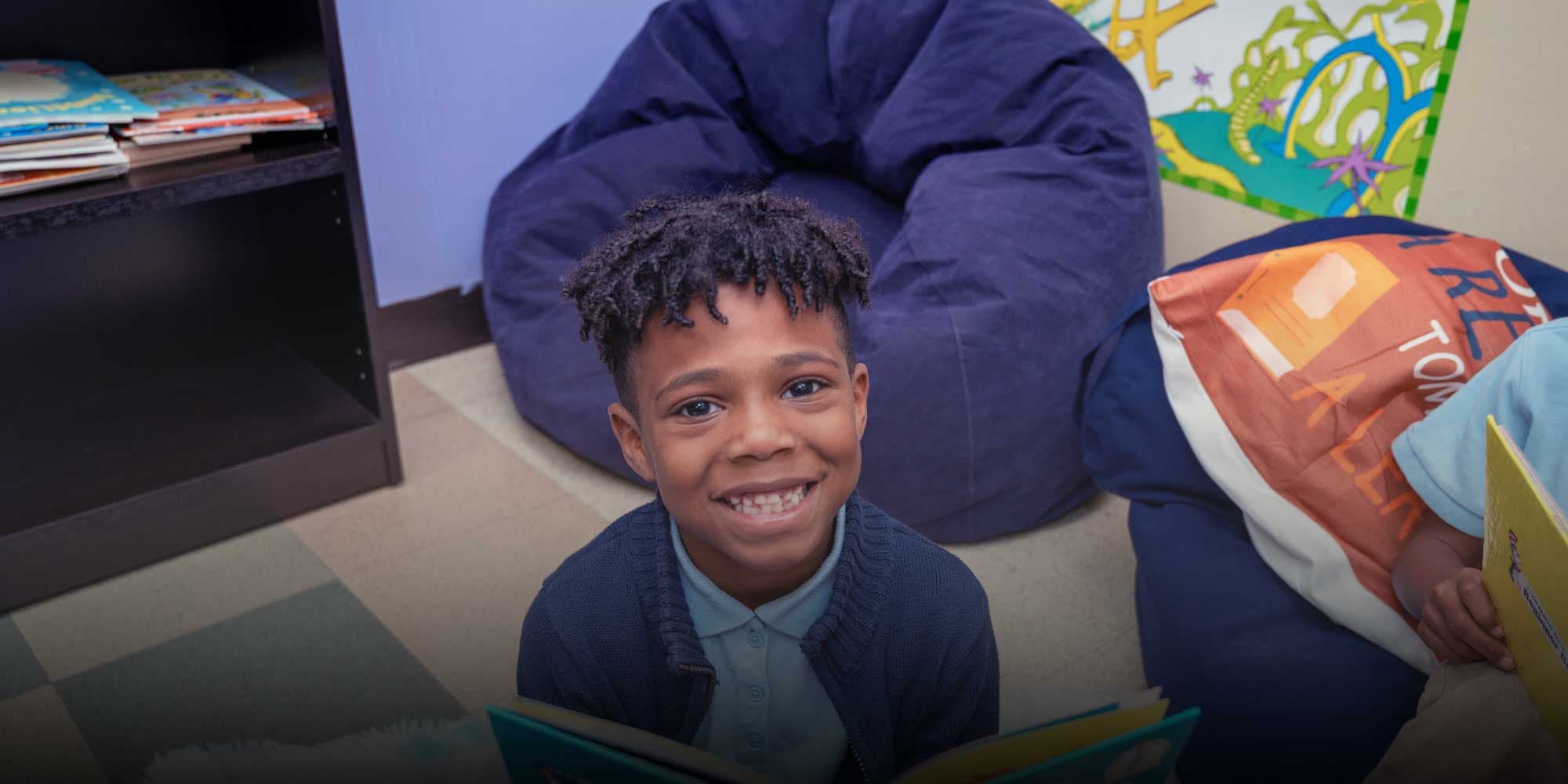 Smiling student holding an open book in a classroom reading corner.