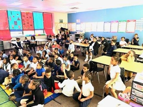 Children sitting on the floor and at desks in a classroom.